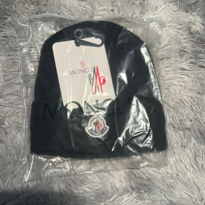 Moncler mössa  - Svart ribbstickad mössa från Moncler med klassisk logotyp framtill. Mössan har en enkel, stilren design och är tillverkad i mjukt material som passar perfekt för kyliga dagar.