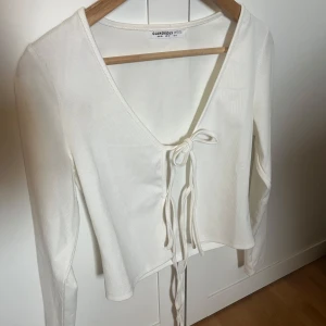 Vit kofta med knyt från Glamorous Petite - Säljer en vit kofta från Glamorous Petite med v-ringning och knyt framtill. Toppen har långa ärmar och är oanvänd. Perfekt för en trendig look.