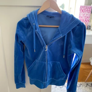 Blå velour hoodie från Juicy Couture - Säljer en snygg blå velour hoodie från Juicy Couture. Den har dragkedja framtill, huva med dragsko och broderad logga på bröstet. Perfekt för en avslappnad stil. 🌀