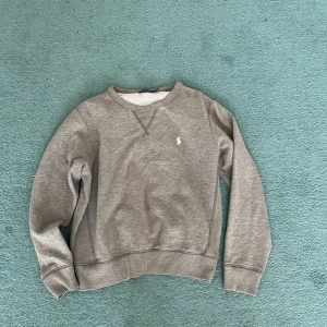 Grå sweatshirt från Polo Ralph Lauren - Säljer en klassisk grå sweatshirt från Polo Ralph Lauren med rund hals och broderad logga på bröstet. Tröjan har långa ärmar och ribbade muddar vid ärmslut och nederkant. Perfekt för en avslappnad stil. Är junior storlek så skulle säga att den passar mellan 160-170 ungefär 
