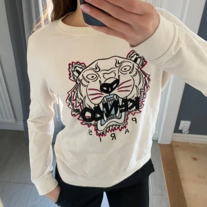 Vit tröja med tigerbroderi från Kenzo - Säljer en vit tröja från Kenzo med ett stort, detaljerat tigerbroderi på framsidan. Tröjan har långa ärmar och en normal passform. Säljer då den tyvärr inte längre kommer till användning 
