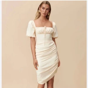 Adoore klänning  - Ljusgul/off white/beige  klänning från Adoore med puffiga kortärmar och en smickrande rynkad design. Klänningen har en fyrkantig urringning och knytning framtill, vilket ger en romantisk touch. Perfekt för speciella tillfällen. Aldrig använd med lapparna kvar. Passar M-L