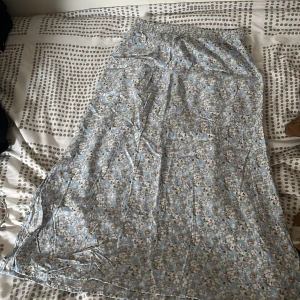 Blommig långkjol i blått och beige - Säljer en söt långkjol med blommigt mönster i blått, beige och vitt. Kjolen har resår i midjan och är tillverkad i ett lätt och luftigt material, perfekt för varmare dagar. Passar dig som gillar en romantisk och avslappnad stil. Kjolen har två korta öppningar längst ner på båda sidorna.