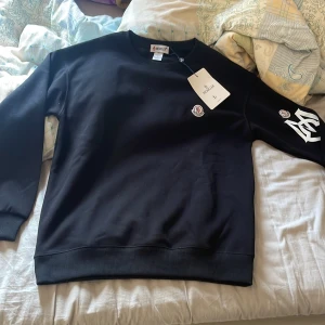 Svart sweatshirt från Moncler - Säljer en svart sweatshirt från Moncler med rund halsringning och vita tryckdetaljer på ärmen. Tröjan har Moncler-logga på bröstet och ärmen. Klassisk och stilren design med ribbade muddar vid ärmslut och nederkant.