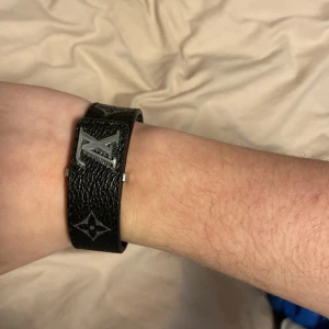 Louis Vuitton svart armband med silverdetaljer - Snyggt svart armband från Louis Vuitton med klassiska LV-loggan och monogramdetaljer i silver. Tillverkat i präglat skinn och har ett stilrent spänne. Perfekt accessoar för att lyfta din outfit. Har gjort några extra hål så det passar vem som helst