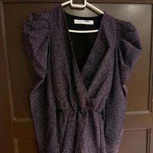 Glittrig lila klänning med puffärmar  - Säljer en glittrig lila klänning/shorts från junkyard med markerade axlar och puffärmar. Klänningen har omlott-v-ringning och resår i midjan för en snygg siluett. Perfekt för festliga tillfällen!