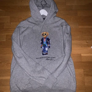 Grå hoodie med Polo Bear från Ralph Lauren - Säljer en grå hoodie från Ralph Lauren med den ikoniska Polo Bear-trycket på bröstet. Tröjan har huva och ribbade muddar vid ärmslut och nederkant. Perfekt för en avslappnad och stilren look.