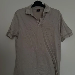 Beige pikétröja från BOSS - Säljer en klassisk beige pikétröja från BOSS med korta ärmar och krage. Tröjan har två knappar vid halsen och diskret BOSS-logga på bröstet. Perfekt för en stilren och avslappnad look.