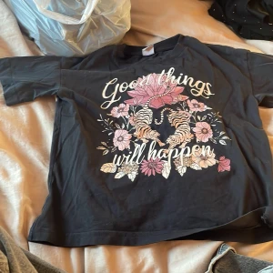 Svart t-shirt med blommigt tryck från Lindex - Säljer en svart t-shirt från Lindex med ett stort blommigt tryck och texten 'Good things will happen' på framsidan. Perfekt för en avslappnad stil.