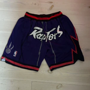 Lila Toronto Raptors shorts från Just Don - Säljer ett par lila Toronto Raptors shorts från Just Don med coolt blixtmönster och lagets logga. Shortsen har en elastisk midja med röd och svart rand. Perfekta för basketfans som vill sticka ut!