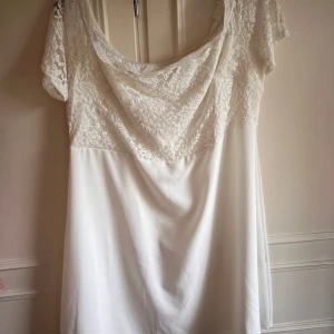 Vit klänning med spetsdetaljer - Elegant vit klänning med spetsdetaljer på överdelen och korta ärmar. Klänningen har en dragkedja på sidan och är tillverkad i polyester med en liten del elastan för extra komfort. Perfekt för sommarens festligheter!