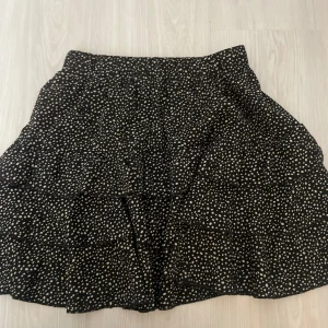 Svart kortkjol med prickigt mönster - Snygg svart kortkjol med vita prickar. Kjolen har en luftig passform och är perfekt för både vardag och festliga tillfällen. Den är gjord av ett lätt och bekvämt material som ger en skön känsla hela dagen.