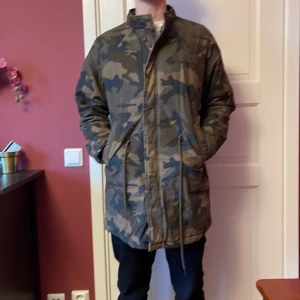 Camouflage parkajacka - Säljer en cool camouflagefärgad parkajacka med dragkedja och snörning i midjan. Jackan har långa ärmar och flera praktiska fickor. Perfekt för en avslappnad stil.