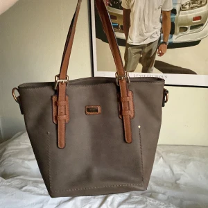 Brun axelväska med detaljer - Snygg brun axelväska med justerbara remmar i en ljusare brun nyans. Väskan har en stilren design med metallspännen och en liten logotyp på framsidan. Perfekt för att bära dina dagliga nödvändigheter med stil.