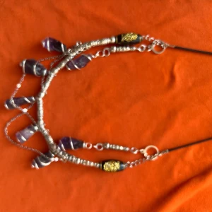 Unikt halsband med lila stenar och silverdetaljer - Säljer ett halsband med flera lila stenar, silverfärgade kedjor och detaljer samt svarta och guldiga inslag. Halsbandet har en bohemisk och iögonfallande design, perfekt för att lyfta din stil.
