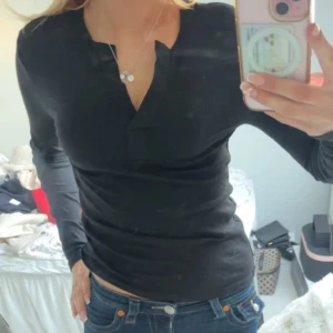 Svart ribbad långärmad topp - Säljer en stilren svart ribbad topp med lång ärm och v-ringning. Perfekt för en enkel och snygg look. Passar bra till jeans eller kjol.