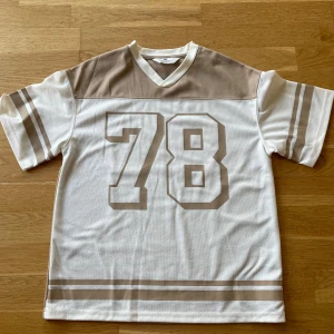 Vit och beige t-shirt med nummer från H&M - Säljer en cool vit och beige t-shirt från H&M med nummer 78 tryckt på både fram- och baksidan. T-shirten har korta ärmar och en v-ringad hals. Perfekt för en avslappnad stil!