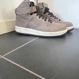 Bruna Nike Air Force i mocka - Säljer ett par bruna Nike Air Force sneakers i mocka. Skorna har en hög modell med snörning och en rem runt ankeln för extra stöd. Den vita sulan ger en snygg kontrast till det bruna mockamaterialet. de är även använda ett fåtal gånger så det är jättebra skick som man ser på bilden