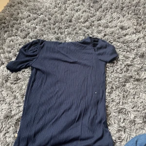 Mörkblå ribbad topp från Pomp de Lux - Mörkblå ribbad topp från Pomp de Lux med korta puffärmar. Enkel och stilren design med rund halsringning. Perfekt för dig som gillar en klassisk look med en twist.