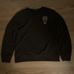 Brun tröja från Morris - Säljer en stilren brun tröja från Morris med ett broderat emblem och brittisk flagga på bröstet. Tröjan har långa ärmar och en klassisk rund halsringning. Perfekt för en avslappnad och snygg look. Storlek: M, Skick: 10/10