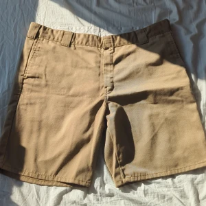 Beige master shorts från Carhartt - Säljer ett par klassiska beige shorts från Carhartt. De har en normal passform och är tillverkade i en blandning av polyester och bomull. Shortsen har hällor för bälte och en bakficka med knapp. Perfekta för en avslappnad stil.