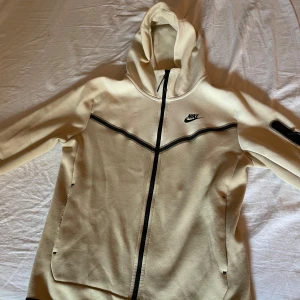 Nike tech hoodie  - Säljer en snygg beige huvtröja från Nike med dragkedja och huva. Tröjan har svarta detaljer och en ficka med dragkedja på ärmen. Perfekt för en sportig look! 