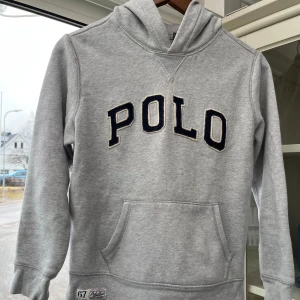 Grå hoodie från Polo Ralph Lauren - Grå hoodie med svart POLO-tryck på bröstet och magficka. Klassisk design med huva och ribbade muddar. Tillverkad i mjuk bomullsblandning för extra komfort. Inga större skador en lappen. Skriv om ni har några funderingar. Jalla mannen🥀🥀🥀