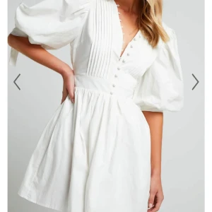 Studentklänning!  - Säljer en elegant vit klänning med puffiga trekvartsärmar och knappar framtill. Klänningen har en v-ringad design och fina veckdetaljer på bröstet. Perfekt för en stilren look! Använd endast en gång på min egna student, hade inte rekommenderat någon annan klänning!! Klänningen är från australiska märket showpoo, köpt för ungefär 1100 och frakt på 600! 