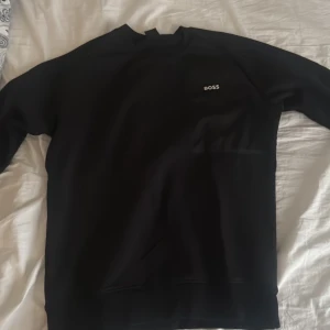 Svart tröja från Boss - Säljer en stilren svart tröja från Boss. Tröjan har en enkel design med långärmade ärmar och en liten logotyp på bröstet. Perfekt för en avslappnad och snygg look.