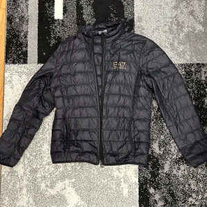 EA7 jacka  - Snygg svart pufferjacka från Emporio Armani med EA7-logga i guld på bröstet. Jackan har en dragkedja framtill och är quiltad för extra värme. Perfekt för kyligare dagar.                             Kategori: höstjacka