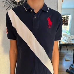 Mörkblå pikétröja från Ralph Lauren - Säljer en snygg mörkblå pikétröja från Ralph Lauren med en vit diagonal rand och röd logga på bröstet. Tröjan har korta ärmar och en amerikansk flagga på ärmen. Perfekt för en stilren look! Den är storlek L/G för barn mellan 14-16 vilket motsvarar typ S i vuxenstorlek!!