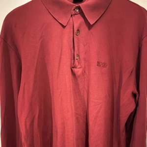 Röd pikétröja från Boss - Snygg röd pikétröja från Boss med klassisk krage och knappar framtill. Perfekt för en stilren look. Tröjan har långa ärmar och är tillverkad i ett mjukt material. (Passar även medium)
