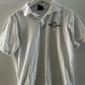 Vit pikétröja från Peak Performance - Säljer en stilren vit pikétröja från Peak Performance med broderad logga på bröstet. Tröjan har en klassisk krage och knappar framtill. Perfekt till sommaren.