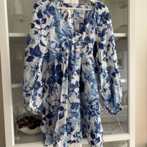 Blåvit blommig klänning - Charmig blåvit klänning med blommigt mönster. Den har en v-ringad design och långa ärmar. Perfekt för en sommaren 