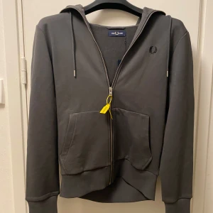 Grå hoodie från Fred Perry - Snygg grå hoodie från Fred Perry med dragkedja och klassisk logga på bröstet.                  Helt ny aldrig andvänd