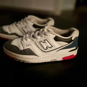 New Balance 550 sneakers - Snygga New Balance 550 sneakers i vitt med grå och blå detaljer. Skorna har en rund tå och snörning, med en kontrasterande röd sula. Perfekta för en stilren och sportig look. Pris kan diskuteras!