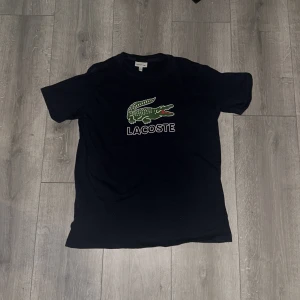 Mörk blå t-shirt från Lacoste - Säljer en svart t-shirt från Lacoste med den ikoniska gröna krokodilloggan och texten 'Lacoste' på bröstet. Perfekt för en stilren och avslappnad look. 🐊