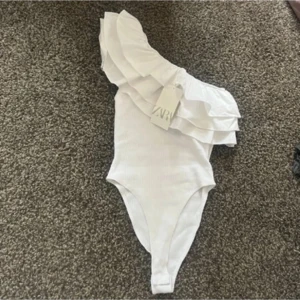 Zara body  - Säljer en stilren vit oneshoulder topp från Zara med volangdetaljer. Perfekt för en trendig look. Toppen är ärmlös och har en elegant passform. Säljer då den är för liten. Bara använd två gånger!