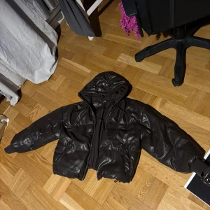 Svart pufferjacka med huva - Säljer en mörkbrun pufferjacka med huva. Jackan har dragkedja framtill och justerbara snören vid nederkanten. Perfekt för kyligare dagar. Den är lite sliten på hängaren annars är den som ny. 