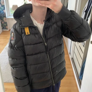 Svart pufferjacka från Parajumpers - Säljer en svart pufferjacka från Parajumpers med huva och dragkedja. Jackan har en gul detalj vid axeln och en broderad logotyp på ärmen. Perfekt för kyliga dagar.