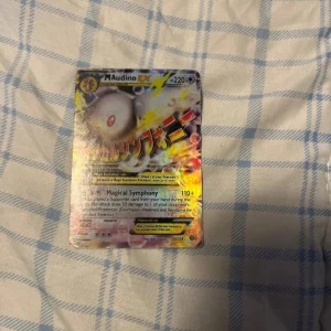 Pokémonkort Mega Audino EX - Säljer ett Pokémonkort med Mega Audino EX. Kortet har en färgglad design med en bild av Mega Audino och text på framsidan. Baksidan har den klassiska blå Pokémon-designen med en Pokéball i mitten. Perfekt för samlare eller spelare som vill förstärka sin kortlek!
