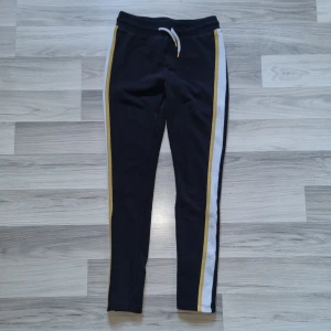 Svarta mjukisbyxor med ränder från H&M - Säljer ett par svarta mjukisbyxor från H&M med vita och gula ränder längs sidorna. Byxorna har en bekväm resårmidja med snörning för justerbar passform. Perfekta för en avslappnad stil.