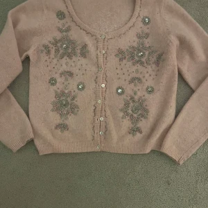 Beige kofta med broderade blommor😍 - Charmig ljus rosa kofta med vackra broderade blommor och pärlor på framsidan och baksidan. Den har långa ärmar och knappar framtill. Perfekt för en elegant och stilren look. Den är liten i storleken 💕