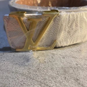 Vit skinnbälte från Louis Vuitton - Säljer ett stilrent vitt skinnbälte från Louis Vuitton med en elegant guldspänne. Perfekt för att ge en lyxig touch till din outfit. Bältet är tillverkat i Spanien och har en klassisk design som passar till många olika stilar.