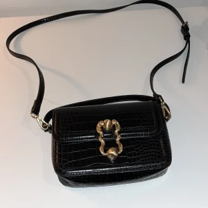 Svart axelväska med gulddetaljer - Snygg svart axelväska från Zara i krokodilskinn (fejk) och med guldfärgad låsdetalj på framsidan. Väskan har en justerbar axelrem. 