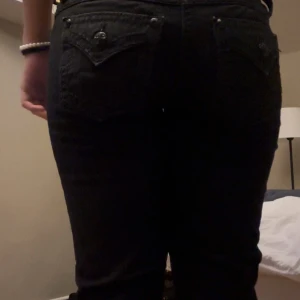 Svarta jeans från Miss Me - Snygga svarta jeans från Miss Me med dekorativa bakfickor och knappar. Jeansen har en normal passform med en lätt modifierad midja och är perfekta för en stilren look. De har en dragkedja och knapp framtill.