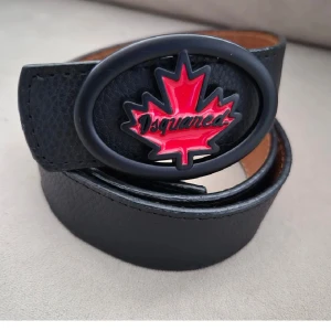 Svart bälte med röd lönnlövsdetalj - Snyggt svart bälte i skinn med en iögonfallande röd lönnlövsdetalj på spännet. Perfekt för att ge din outfit en cool touch. Bältet har en oval form på spännet och flera hål för justering.