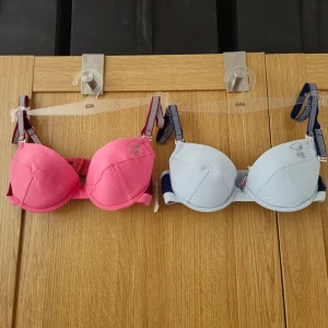 Rosa och blå BH med Snoopy-motiv 65AA - Två söta BH:ar i rosa och blått med ett charmigt Snoopy-motiv. De har glittriga axelband som ger en extra touch av 90-tals stil. Perfekt för den som älskar en Snobben och en lekfull och bekväm stil. Nya, oanvända med prislappen kvar.