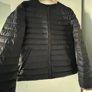 Svart dunjacka från Tommy Hilfiger - Snygg svart dunjacka från Tommy Hilfiger med knappar framtill. Jackan har en stilren design med långa ärmar och diskret logga på bröstet. Perfekt för kyligare dagar.