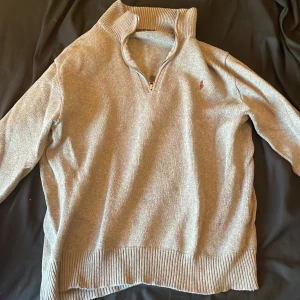 Grå tröja med dragkedja från Ralph Lauren - Säljer en stilren grå tröja från Ralph Lauren med dragkedja och ribbade detaljer vid krage, ärmslut och nederkant. Perfekt för en avslappnad och snygg look.